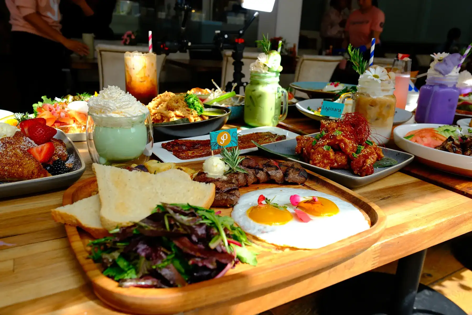 A wooden table crowded with brunch plates, a breakfast board with a fried egg and tall iced drinks